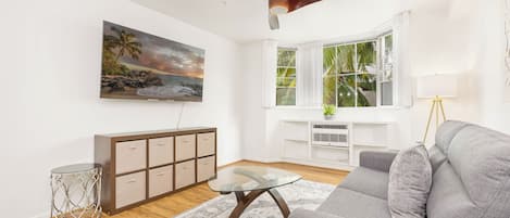 Ett vardagsrum med en grå soffa, ett soffbord i glas, en tavla som föreställer en strand, en takfläkt, ett fönster med vita gardiner och ett litet sidobord.
