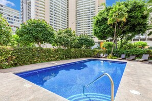 A swimming pool with a metal ladder, surrounded by greenery and tall buildings. - Modern Waikiki 1BR Condo at The Cove w/ Parking/prime Location Near Beach (Honolulu)