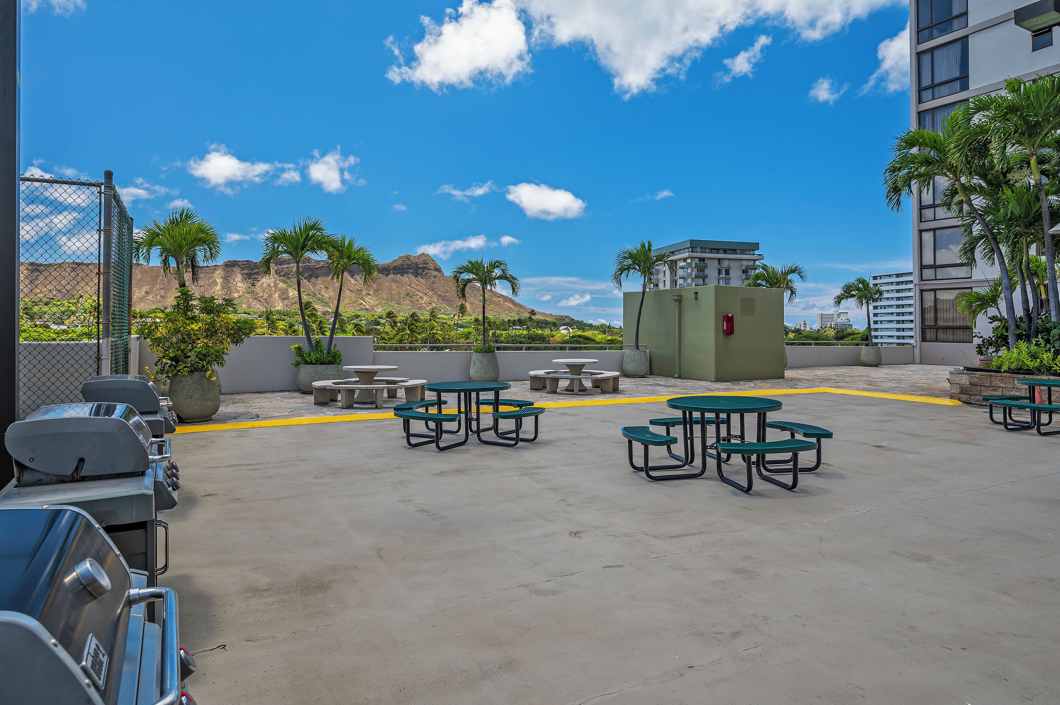 An outdoor patio area with concrete flooring, picnic tables, and a view of a mountain and palm trees.
