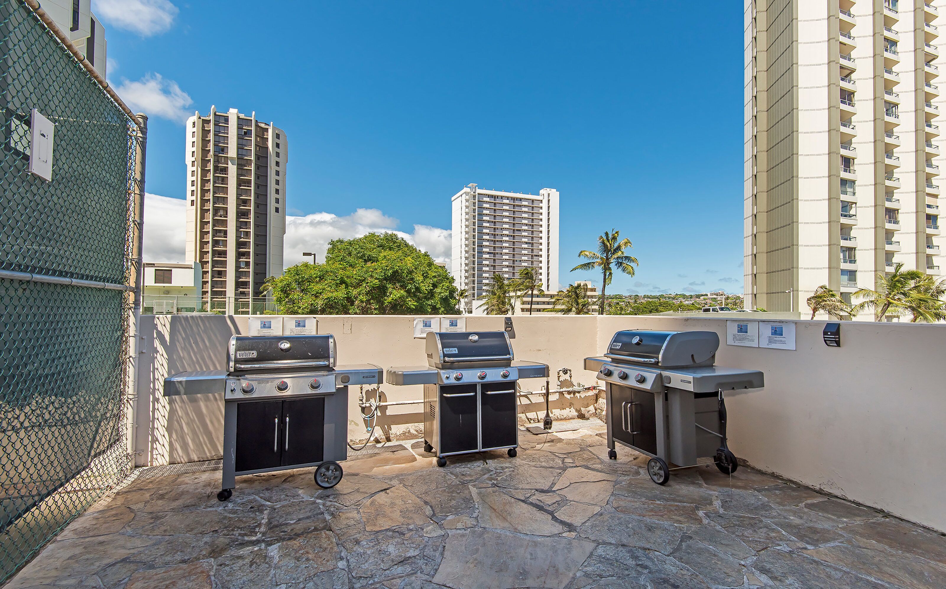 An outdoor patio area with three barbecue grills, a stone-tiled floor, and a view of tall buildings and palm trees in the background.