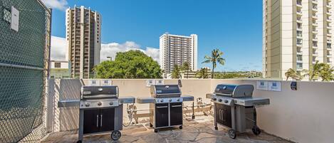 An outdoor patio area with three barbecue grills, a stone-tiled floor, and a view of tall buildings and palm trees in the background.