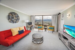A modern living room with a sofa, round coffee table, and a flat-screen TV. A large window offers a city view. - Golf Course-mountain Views Condo at Waikiki Sunset Half a Block to the Beach (Honolulu)