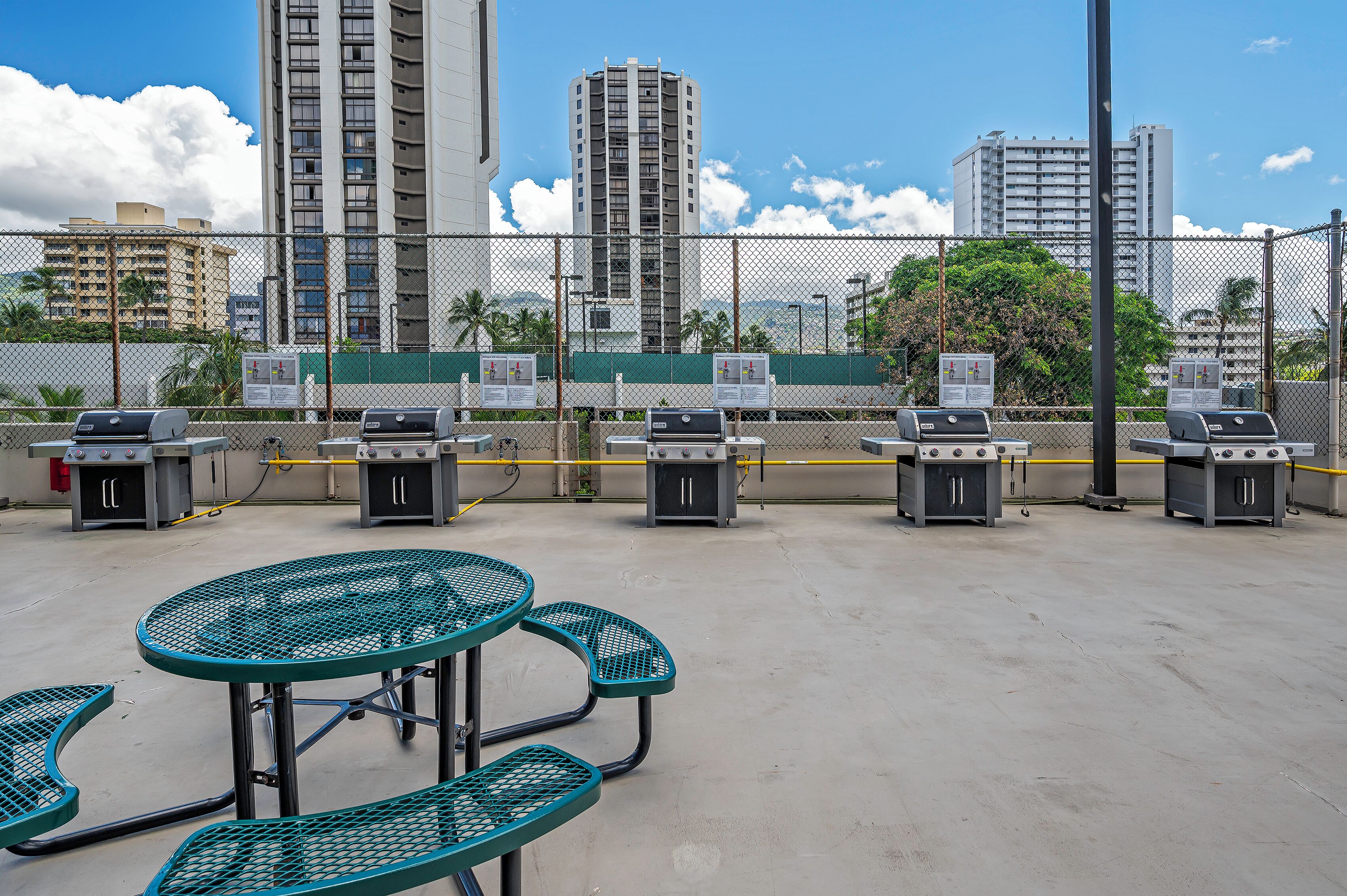 A rooftop patio with outdoor grills, tables, and chairs, surrounded by a chain-link fence.
