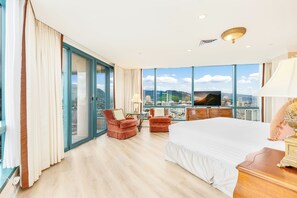 Ein Hotelzimmer mit einem großen Bett, zwei Sesseln, einem Fernseher und Blick auf die Stadt.
