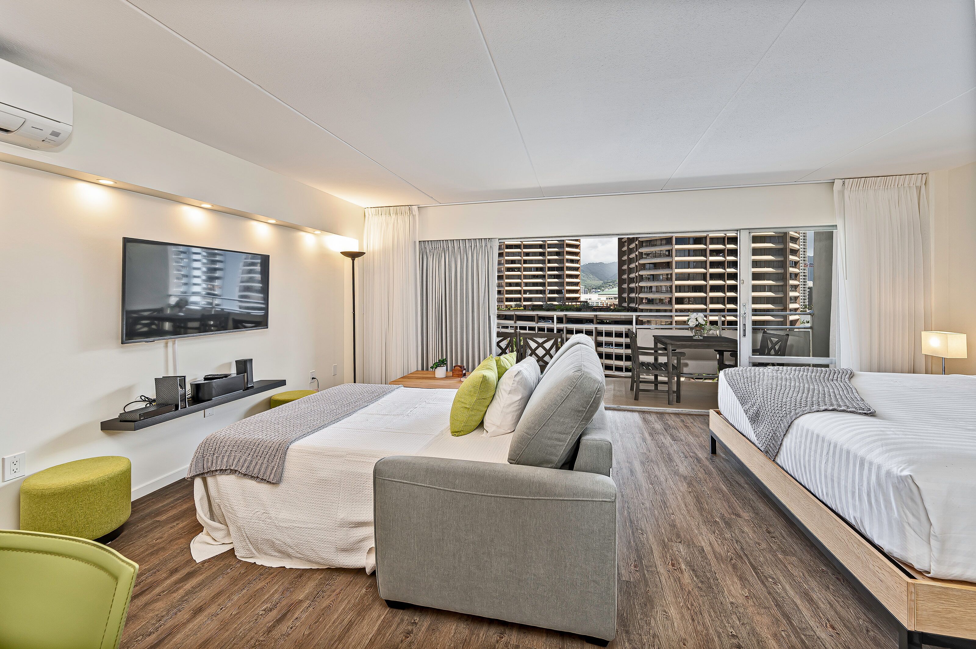 Habitación de hotel con dos camas, un sofá, televisión y balcón con vista a la ciudad.