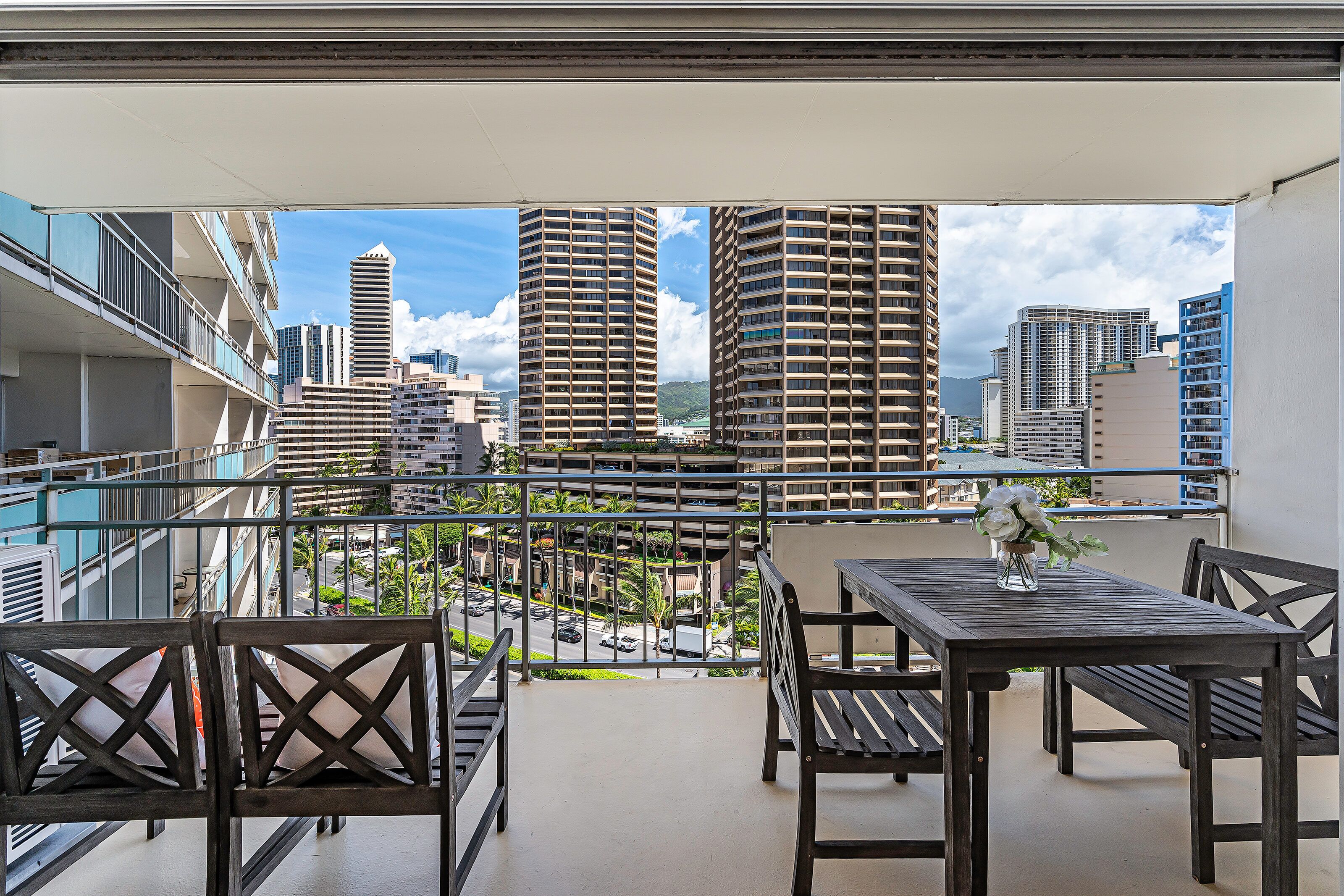 A balcony with a table and chairs, overlooking a cityscape with tall buildings and a clear sky.