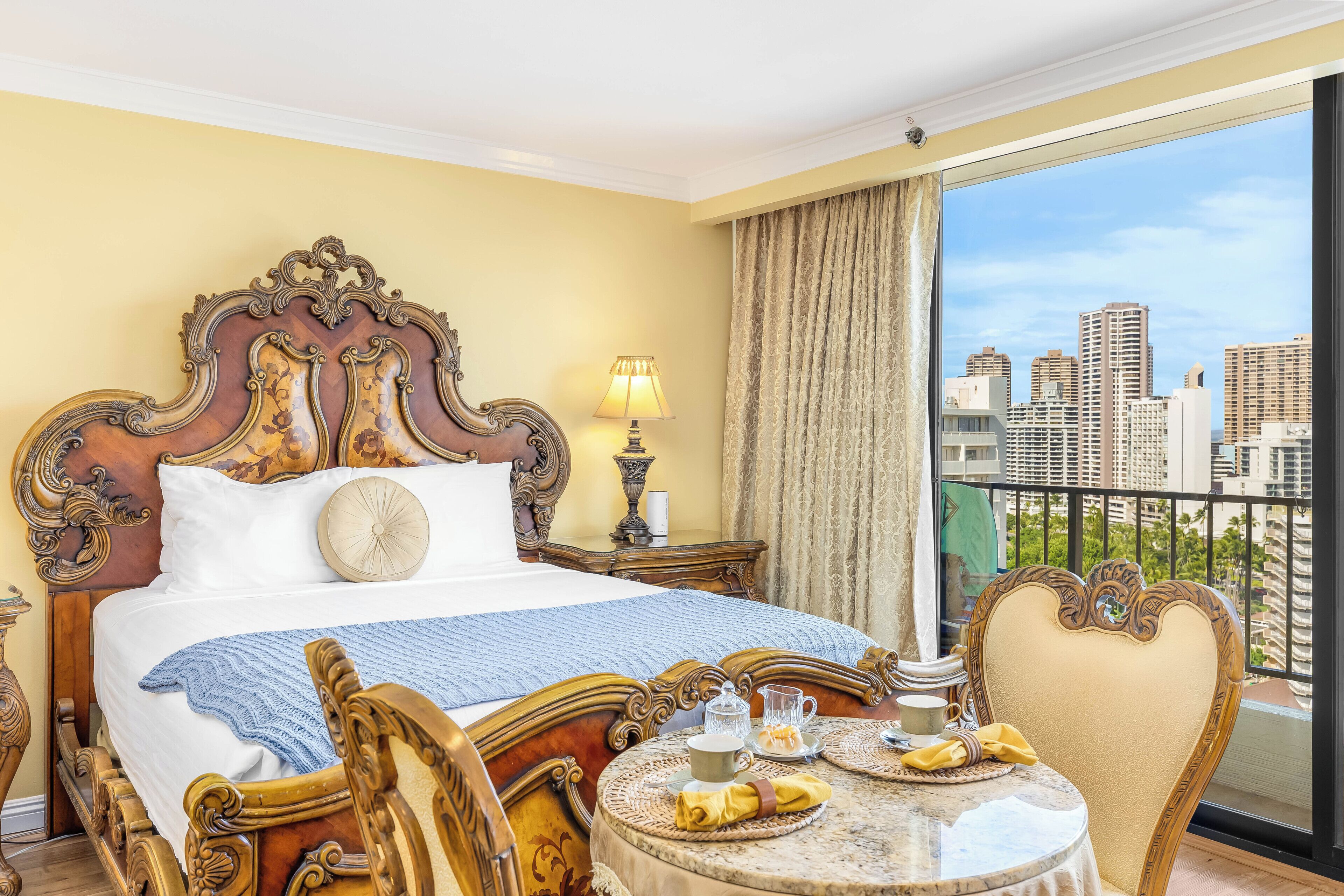 Habitación de hotel con una cama grande, una mesa redonda con alimentos para el desayuno y vistas a edificios de la ciudad.