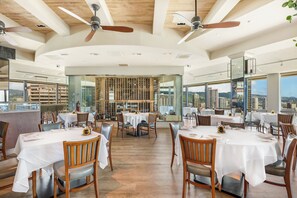 Un restaurant spacieux avec de hauts plafonds, des poutres en bois et de grandes fenêtres offrant une vue sur la ville. Les tables sont dressées avec des nappes blanches et des chaises.