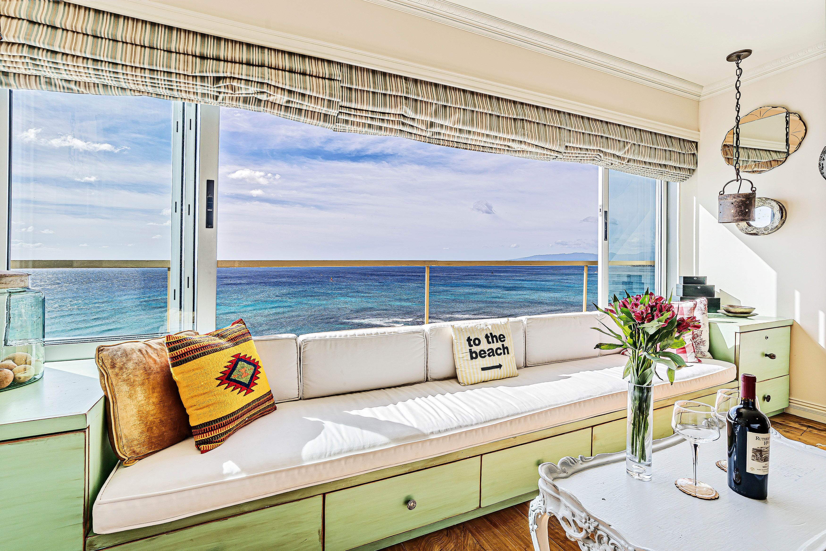 Ein Zimmer mit großem Fenster mit Blick auf das Meer, eine Couch mit dekorativen Kissen, ein kleiner Tisch mit Flasche und Gläsern sowie ein grüner Schrank.