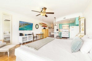 A well-lit bedroom with a large bed, a ceiling fan, a painting of a beach, and a kitchen area with teal cabinets. - Oceanfront W/sunset Views Condo at the Colony Surf Full Kitchen and Parking (Honolulu)
