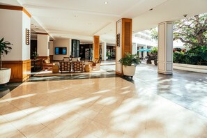 Eine großzügige Lobby mit modernem Design, inklusive einer zentralen Sitzecke mit Sofas und einem Couchtisch, umgeben von großen Kübelpflanzen und einer imposanten Steinskulptur.