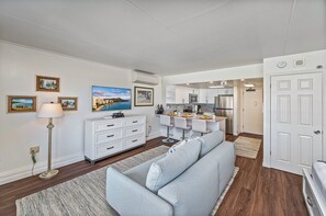 Ein modernes Wohnzimmer mit einem grauen Sofa, einer weißen Kommode und einem Flachbildfernseher. Im Hintergrund ist eine Küche mit weißen Schränken und Edelstahlgeräten erkennbar.