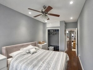 Un dormitorio con cama, mesitas de noche, ventilador de techo, vestidor y baño visible a través de una puerta abierta.