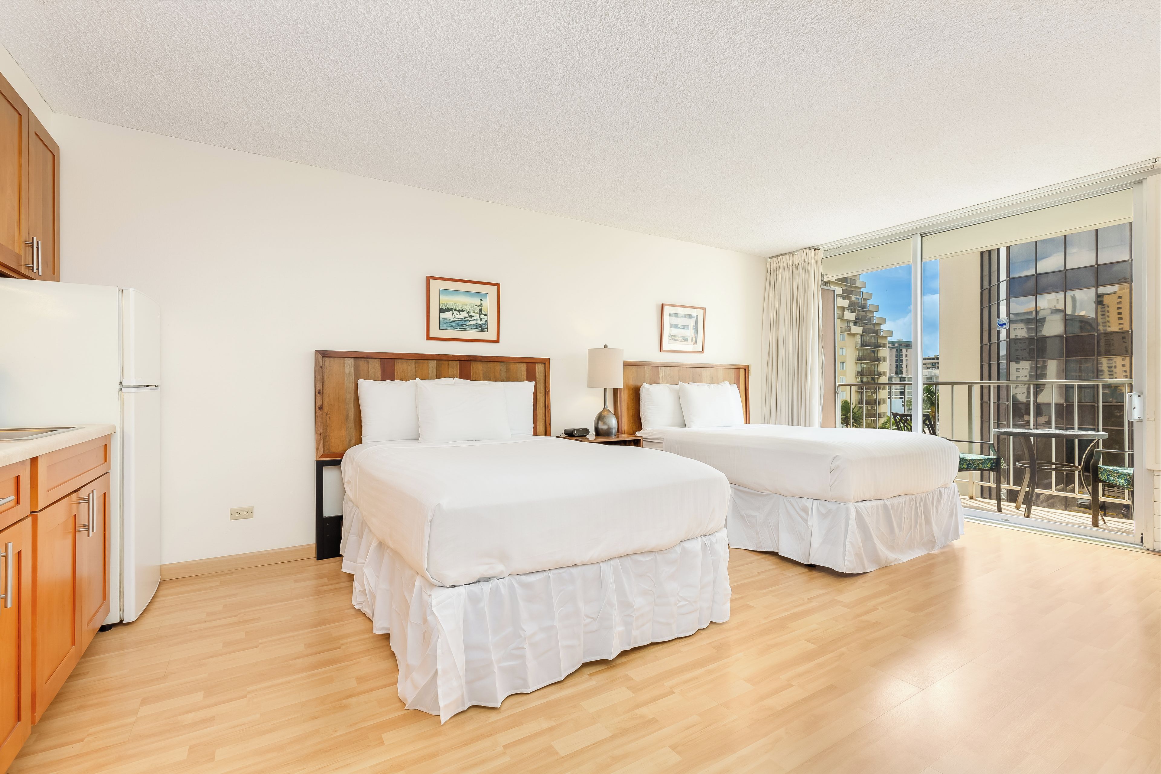 A hotel room with two beds, a kitchenette, and a balcony with a city view.