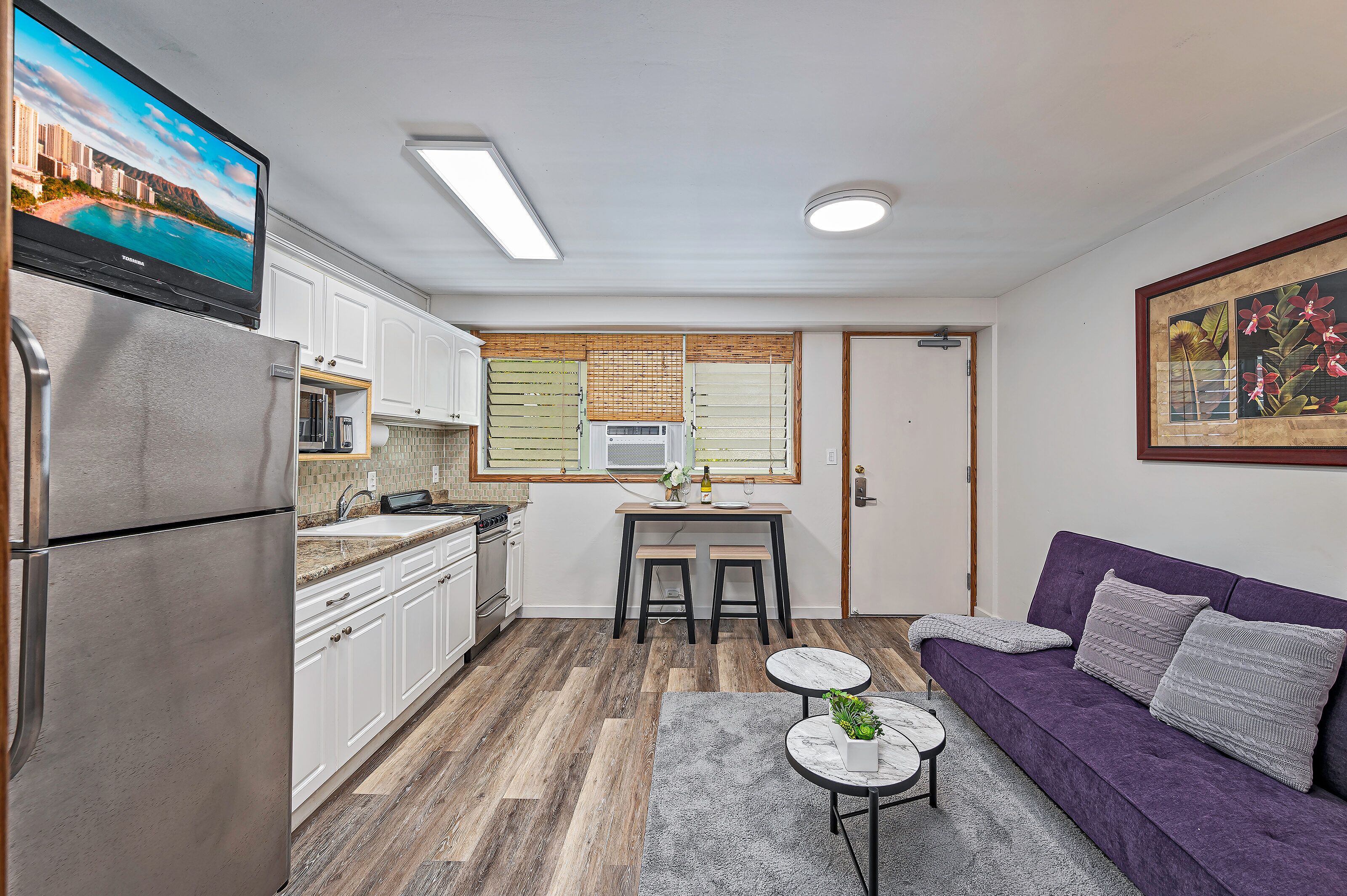 A compact kitchen with a refrigerator, microwave, and stove, a dining area with a table and chairs, and a purple sofa with a gray rug.