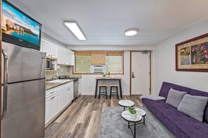 Une cuisine compacte équipée d’un réfrigérateur, d’un four à micro-ondes et d’une cuisinière, un coin repas avec une table et des chaises, et un canapé violet agrémenté d’un tapis gris.
