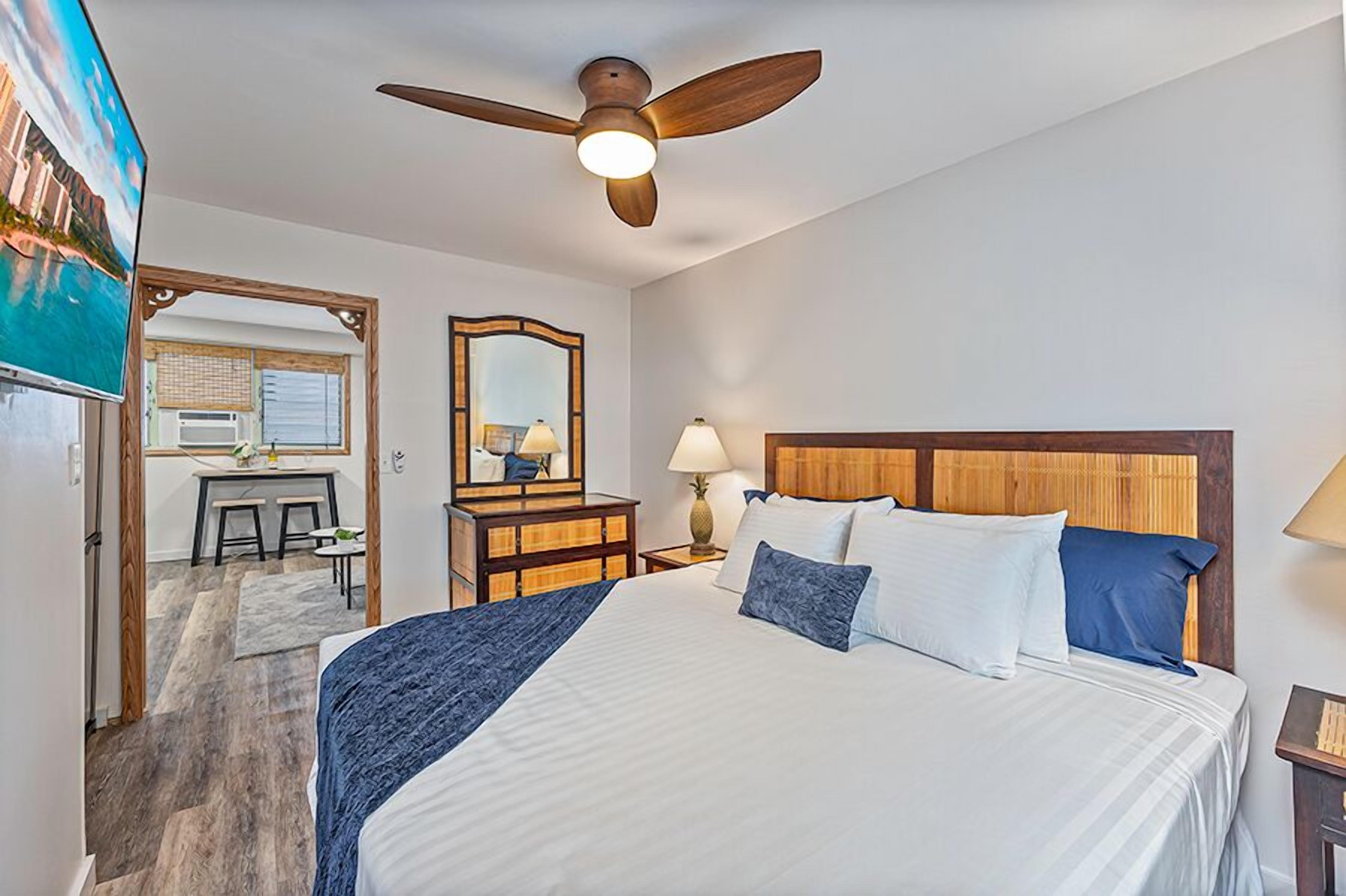 Une chambre d’hôtel avec un lit, un ventilateur de plafond, un bureau avec une chaise, une lampe et un grand miroir.