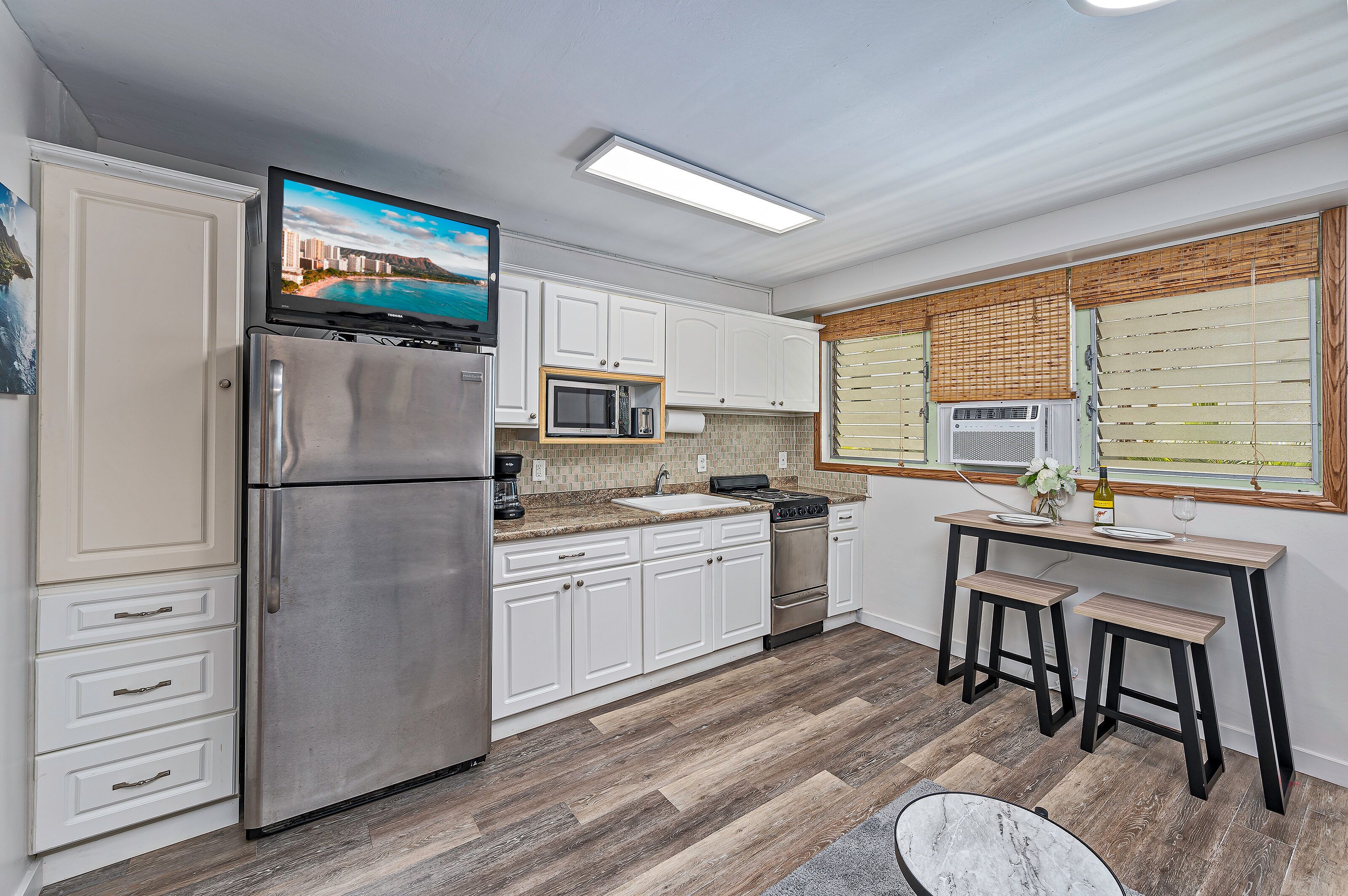 A compact kitchen with white cabinetry, a stainless steel refrigerator, a microwave, and a stove. There is a small dining area with stools and a window with blinds.