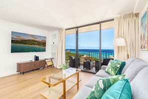 Ein modernes Wohnzimmer mit Sofa, Couchtisch und Fernseher, mit Blick auf einen Balkon und Meer sowie Gebäude.