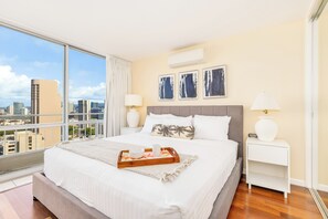 Una camera d'albergo con un letto grande, vista sulla città, un comodino bianco e una lampada.