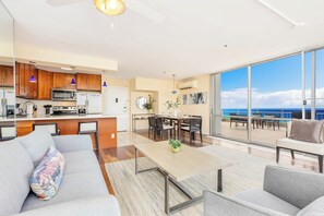 A modern living room with a sofa, coffee table, and dining area. A kitchen with wooden cabinets and appliances is visible. Large windows offer a view of the sky and clouds. - Ocean-marina Views PH at Ilikai Hotel by the Beach W/full Kitchen & Free Wifi (Honolulu)