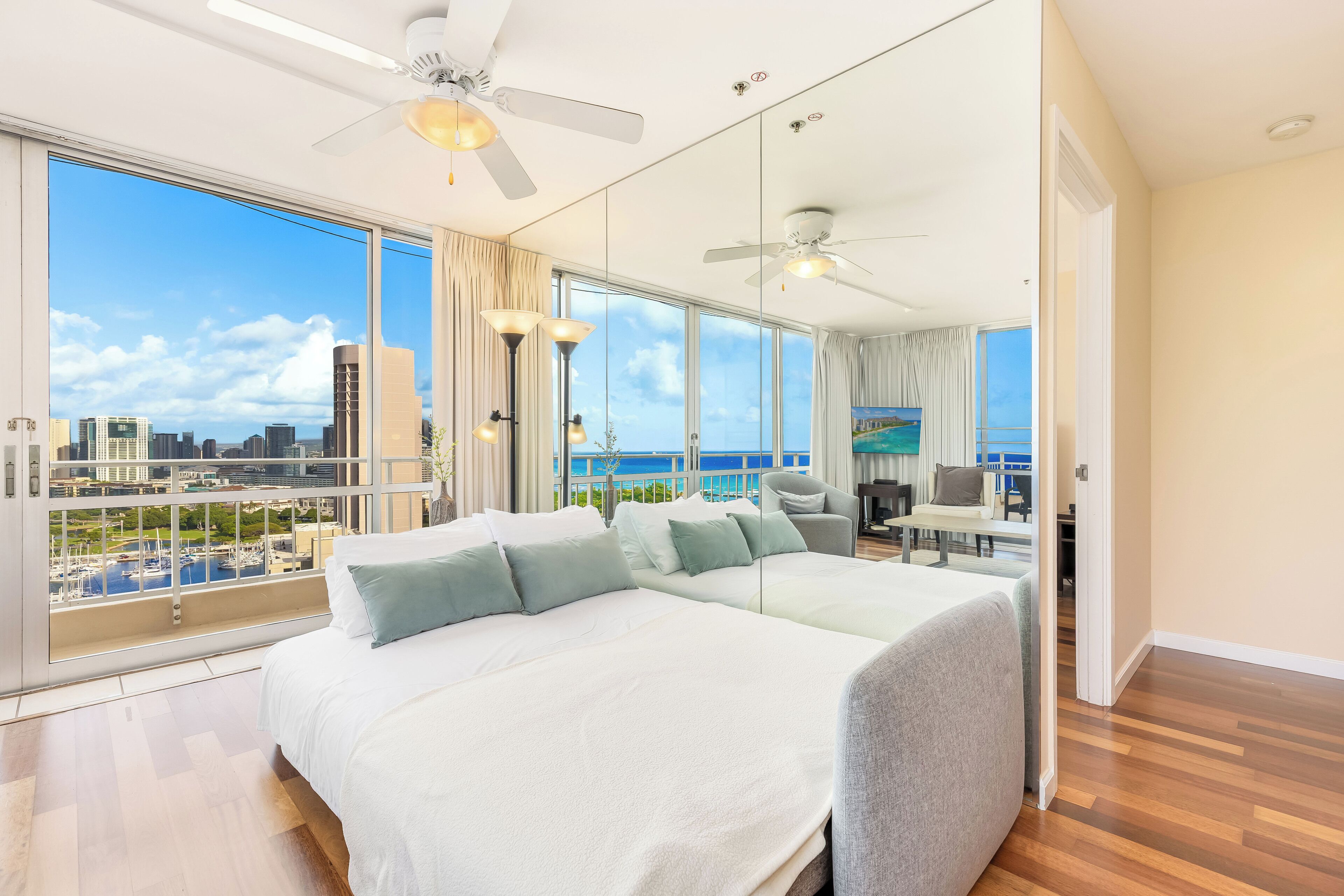 A spacious bedroom with a large bed, a ceiling fan, and a city view through floor-to-ceiling windows.
