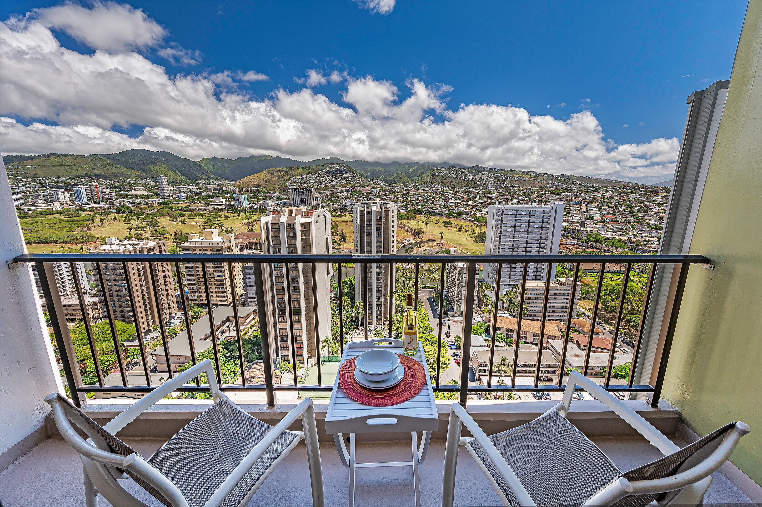 Un balcon avec une table et des chaises, donnant sur un paysage urbain avec des bâtiments et des montagnes.