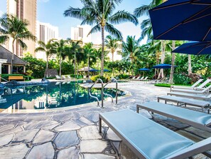 A pool area with lounge chairs and umbrellas, surrounded by palm trees and a cityscape in the background. - Ocean-city Views Glamorous Condo at The Watermark W/parking & Full Kitchen (Honolulu)