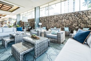 Un espace intérieur vaste et lumineux, aménagé avec des meubles en osier, un mur en pierre et de grandes fenêtres.
