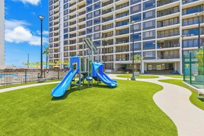 A playground with slides and climbing structures, surrounded by a well-maintained lawn and a large building with multiple balconies. - Mountain and City Tropical Condo at Waikiki Banyan Half a Block to the Beach (Honolulu)