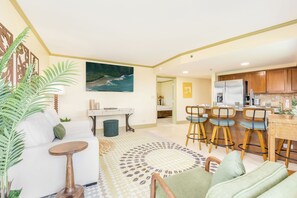 Ein Wohnzimmer mit Sofa, Couchtisch und Sesseln, eine Küche mit Holzausstattung und Kühlschrank, sowie ein Gemälde an der Wand.
