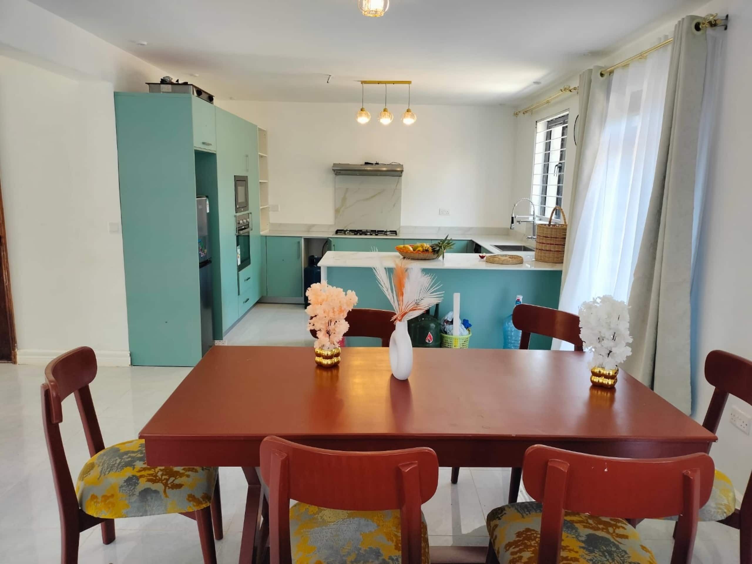 Un'area pranzo con un tavolo e sedie di legno, una cucina sullo sfondo e una finestra con tende.