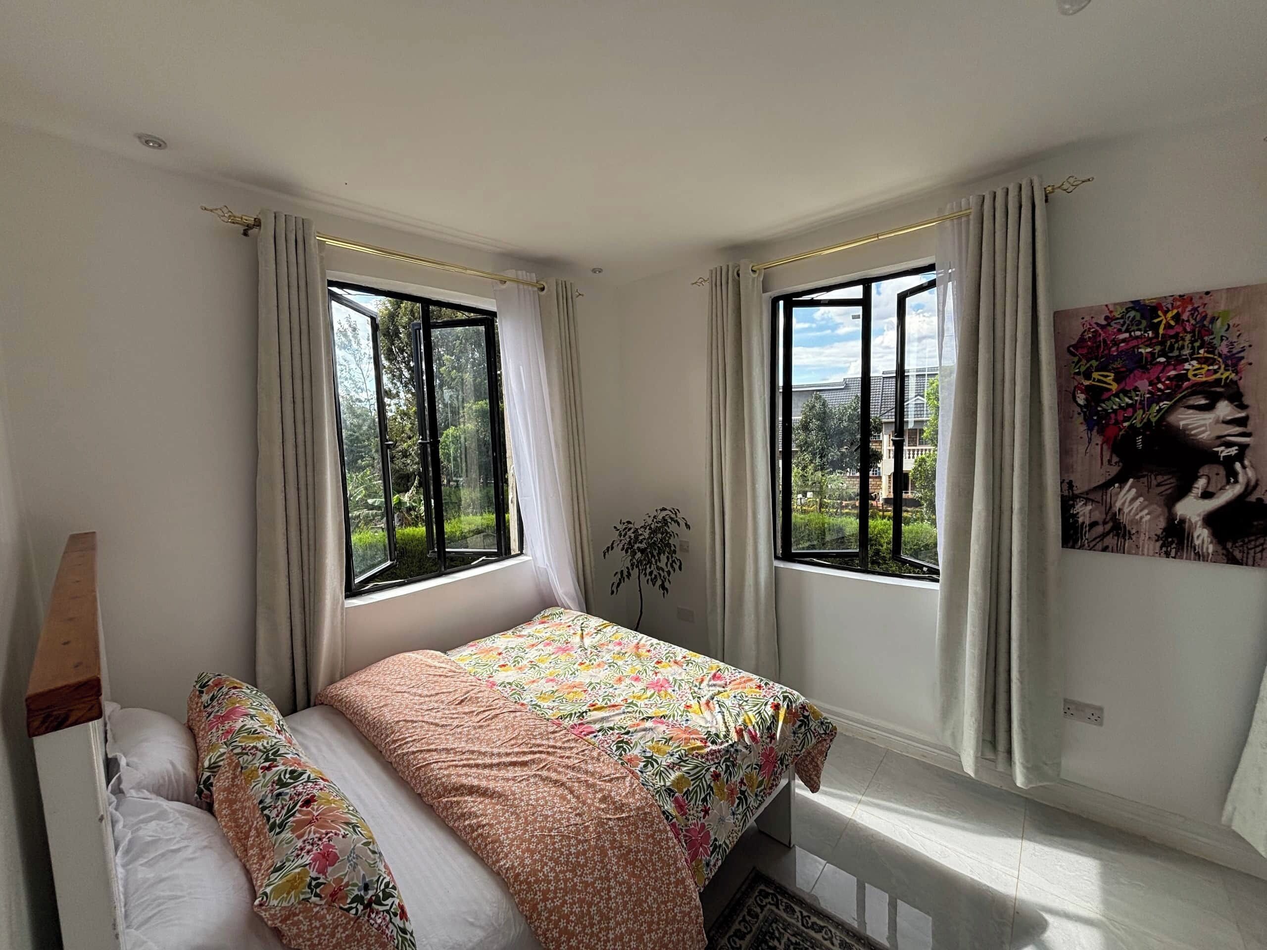 A bedroom with a bed, two windows, a plant, and a painting.