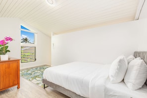 Quarto com cama, penteadeira de madeira, janela com persianas e vista para palmeiras.