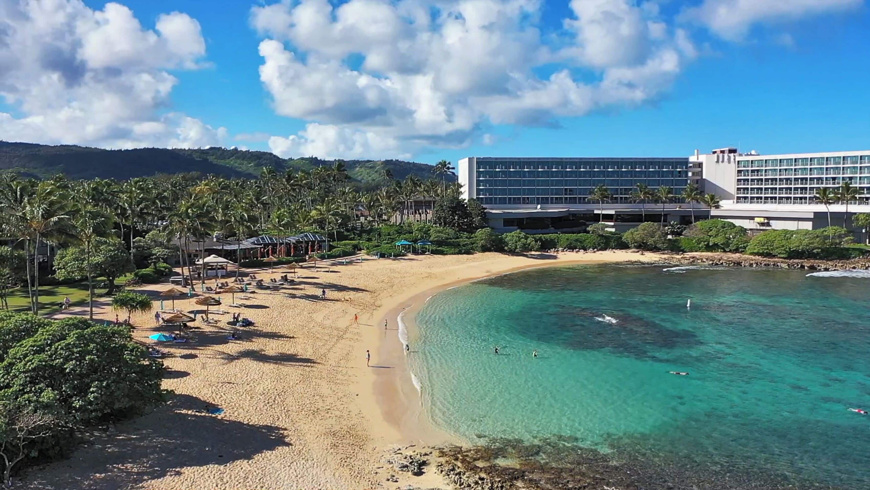 A beach resort with a sandy shore, clear turquoise water, and a large hotel building with multiple floors.