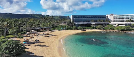 A beach resort with a sandy shore, clear turquoise water, and a large hotel building with multiple floors.