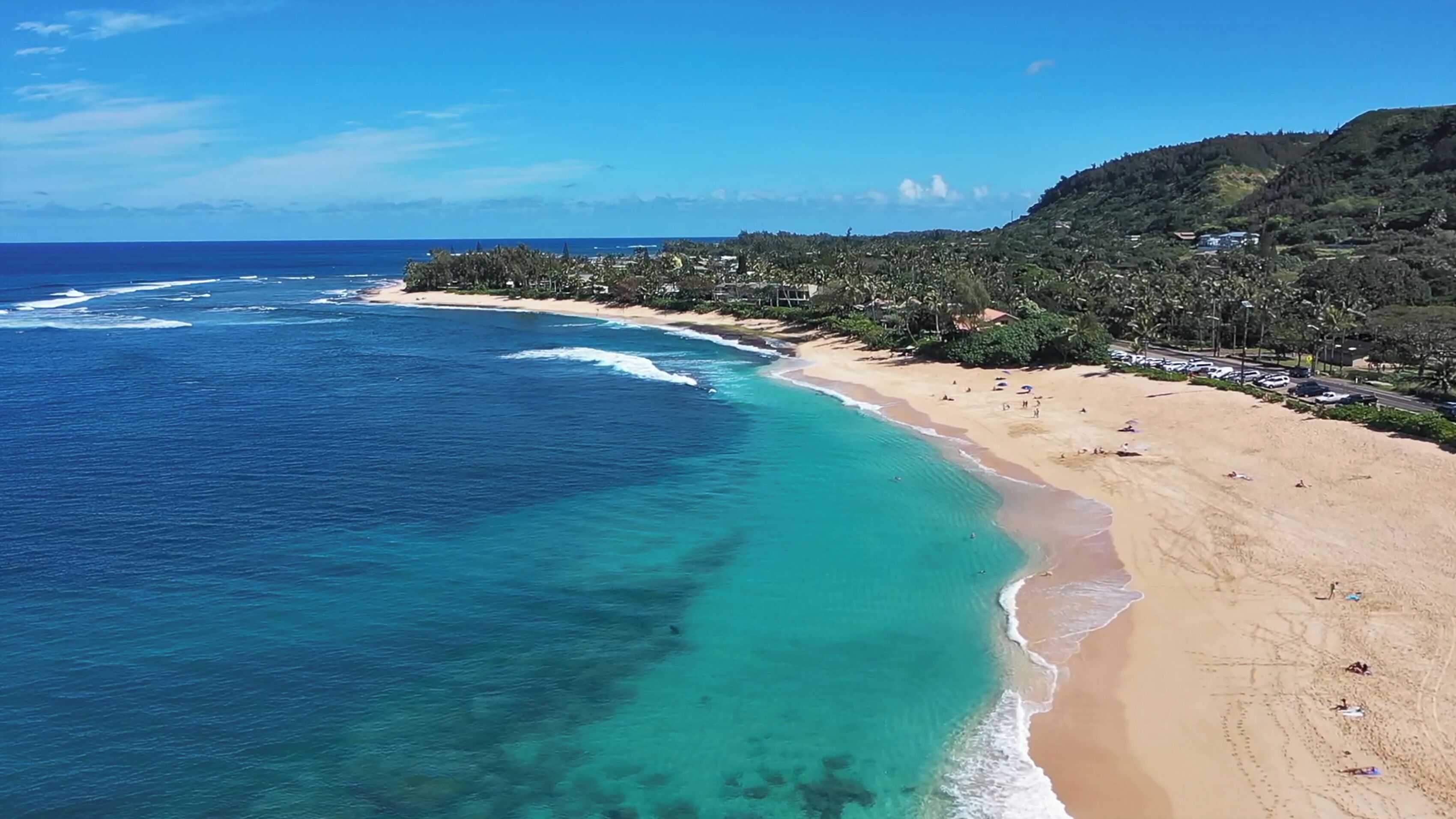 Une plage aux eaux turquoise et limpides, un rivage sablonneux et, en arrière-plan, des collines couvertes d’une végétation luxuriante.