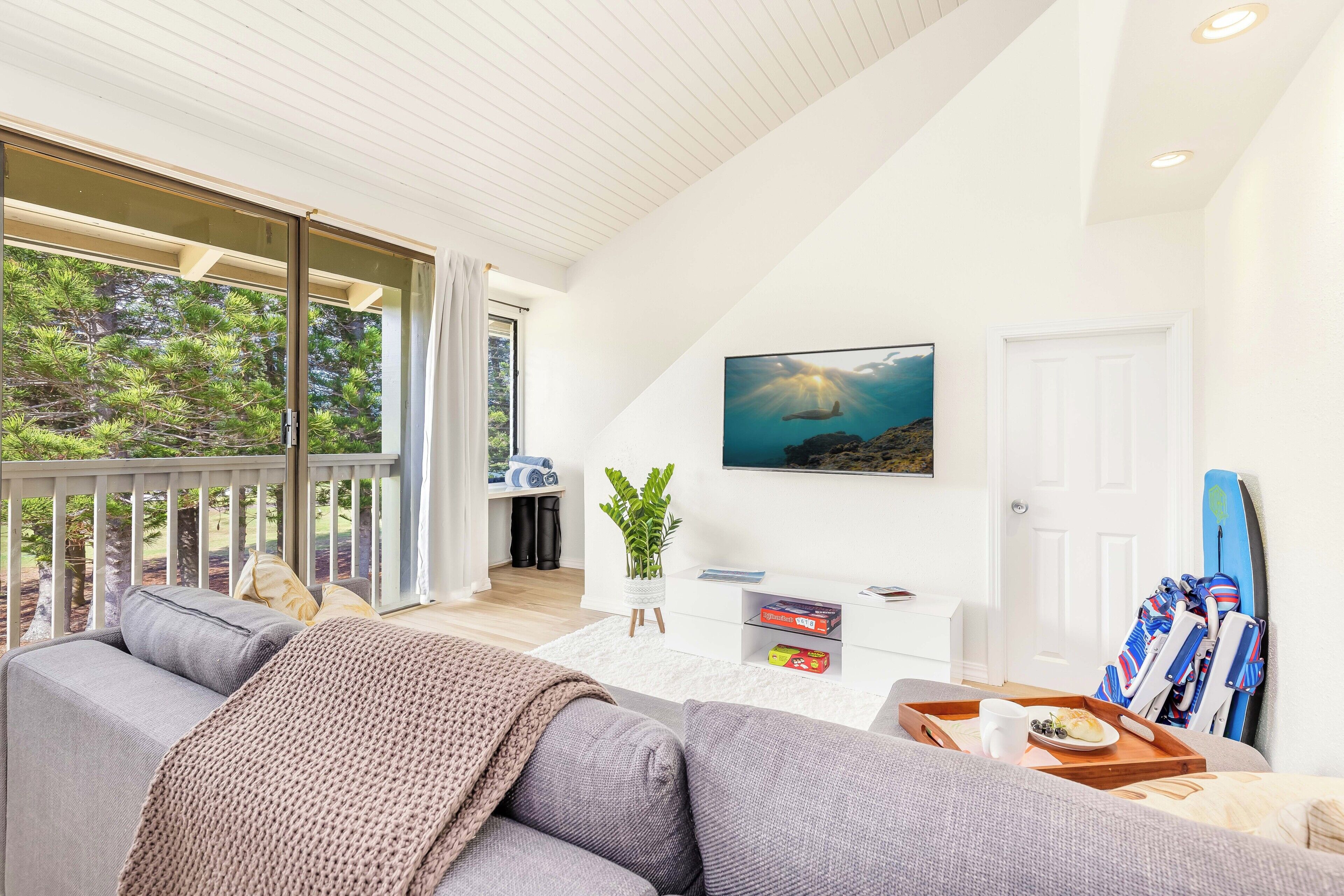 A modern living room with a grey sofa, a white coffee table with a tray of food, a potted plant, and a surfboard leaning against the wall.