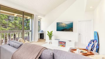 A modern living room with a grey sofa, a white coffee table with a tray of food, a potted plant, and a surfboard leaning against the wall.