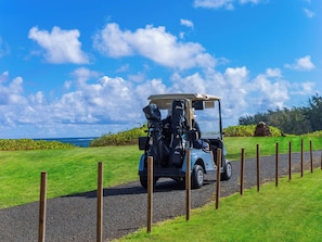 Um carrinho de golfe em um caminho de cascalho, com vista para o oceano e céu claro.