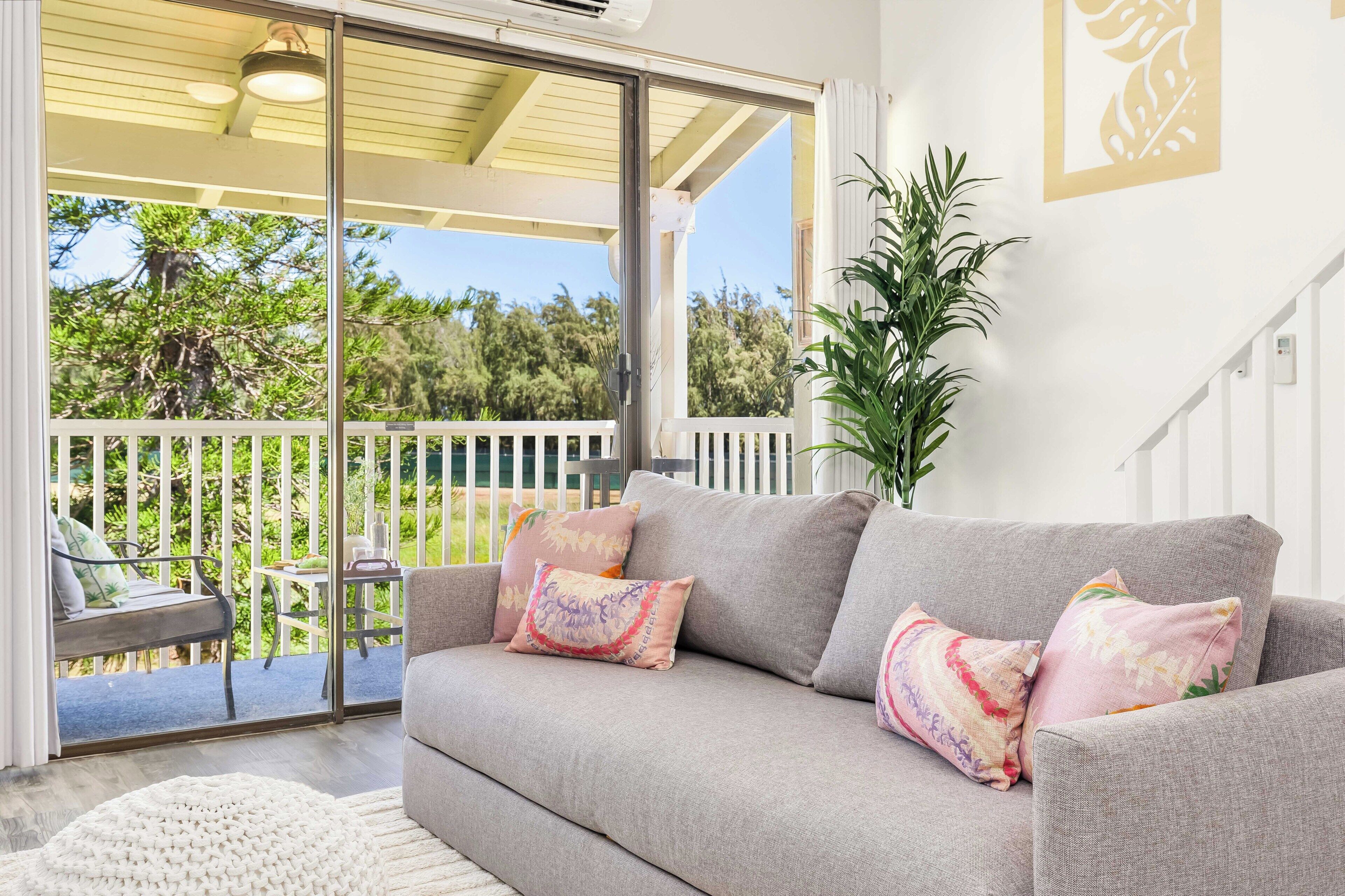 A grey sofa with colorful cushions, a white rug, and a view of a balcony with a table and chairs.