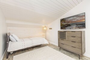 Quarto com cama, penteadeira, pintura de uma praia e teto com ripas de madeira.