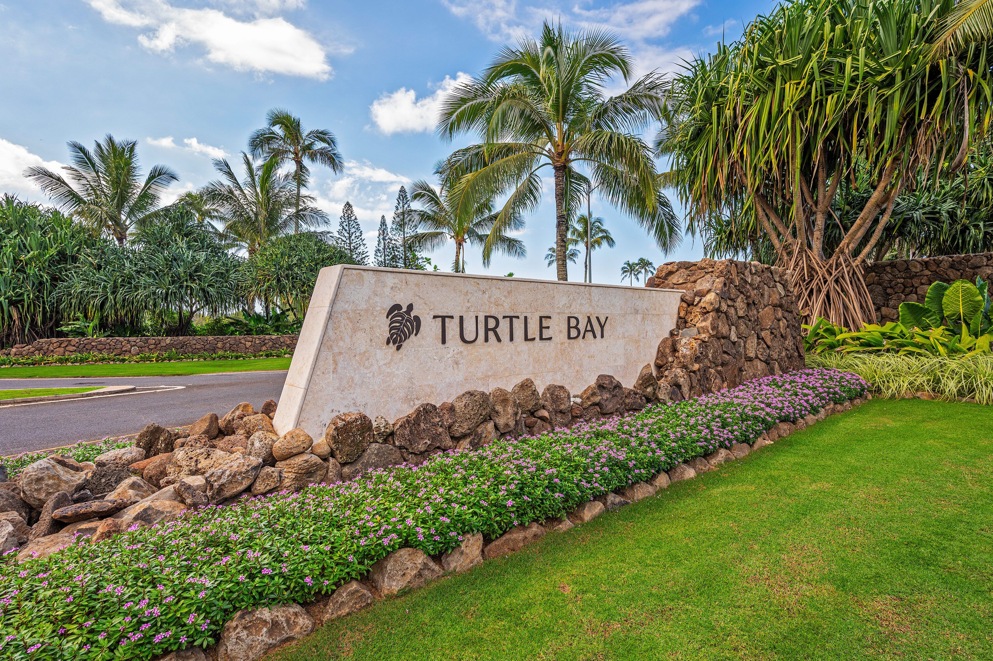 A sign for Turtle Bay with a stone wall, green grass, and tropical plants.