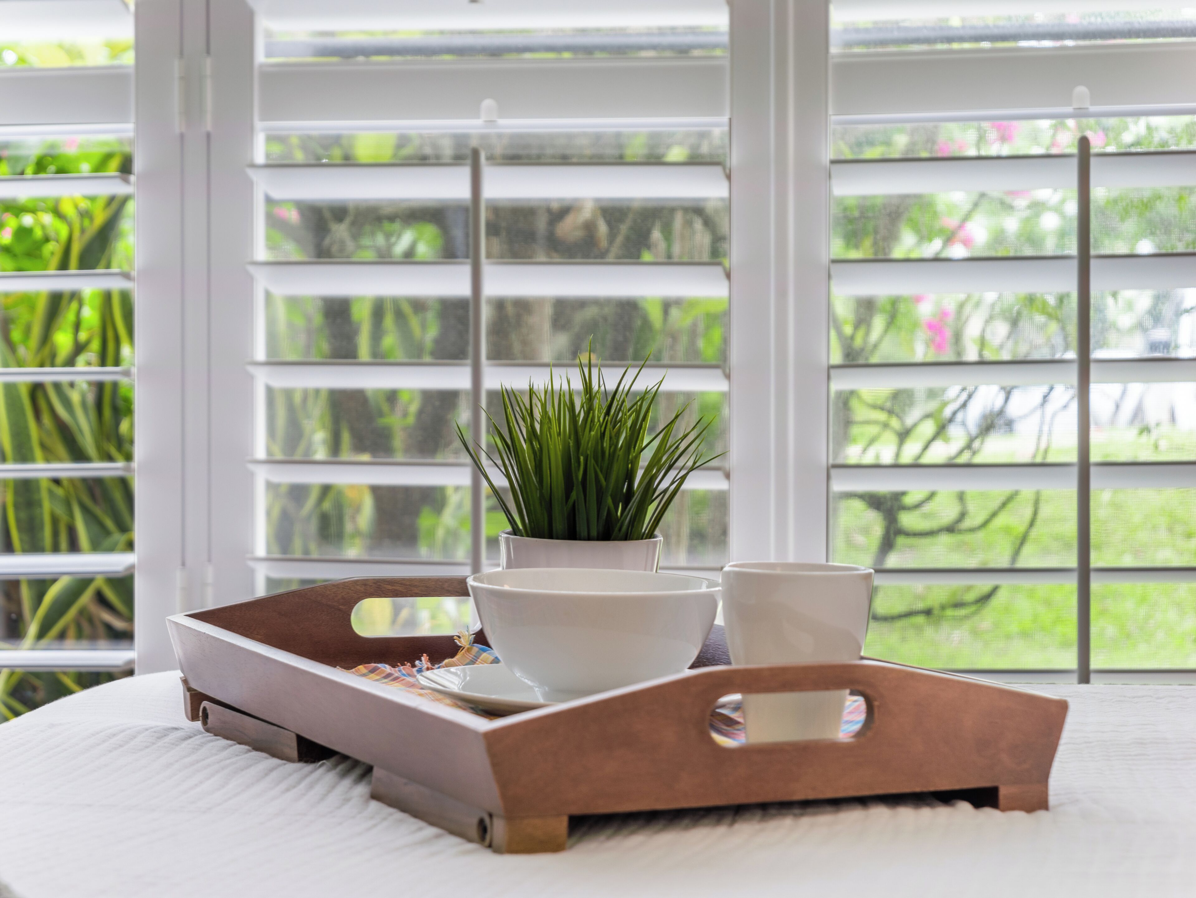 A tray with a potted plant, bowls, and cups on a bed with a view of greenery through a window with blinds.