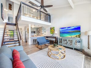 A spacious living area with a high ceiling, a staircase, a kitchen, and a large TV screen. - Exclusive Golf Course Front Kuilima West by Beach W/parking & Great Amenities (Kahuku)