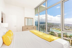 Ein Hotelzimmer mit großem Fenster, einem Bett mit weißer Bettwäsche, einem gelben Kissen und einer Holzkommode.