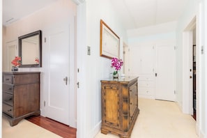 A hallway with a wooden dresser, a mirror, and a vase with flowers. There are white doors and a white wall.
