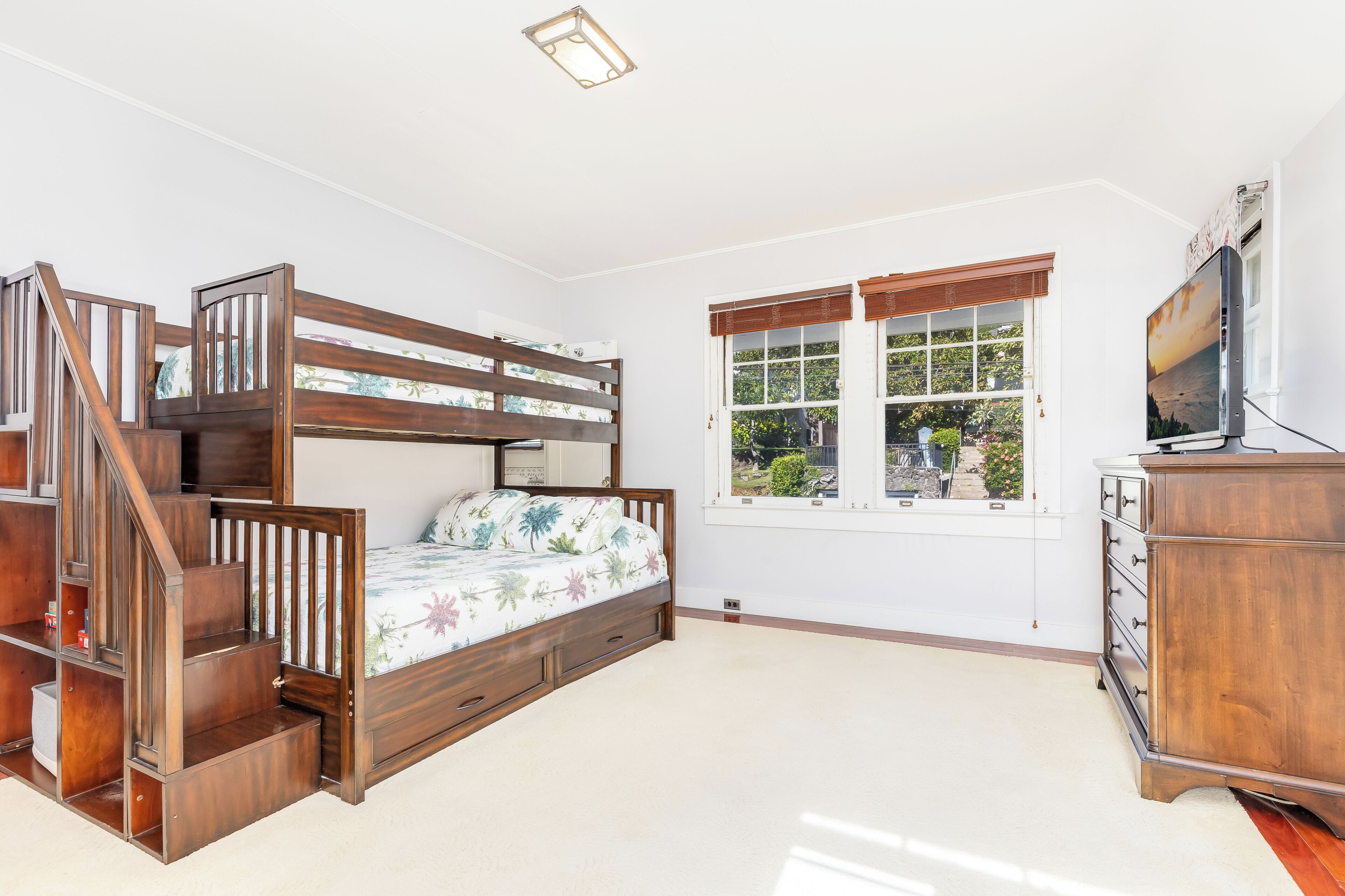 A bedroom with a bunk bed, a wooden dresser, a television, and a window with a view of greenery.