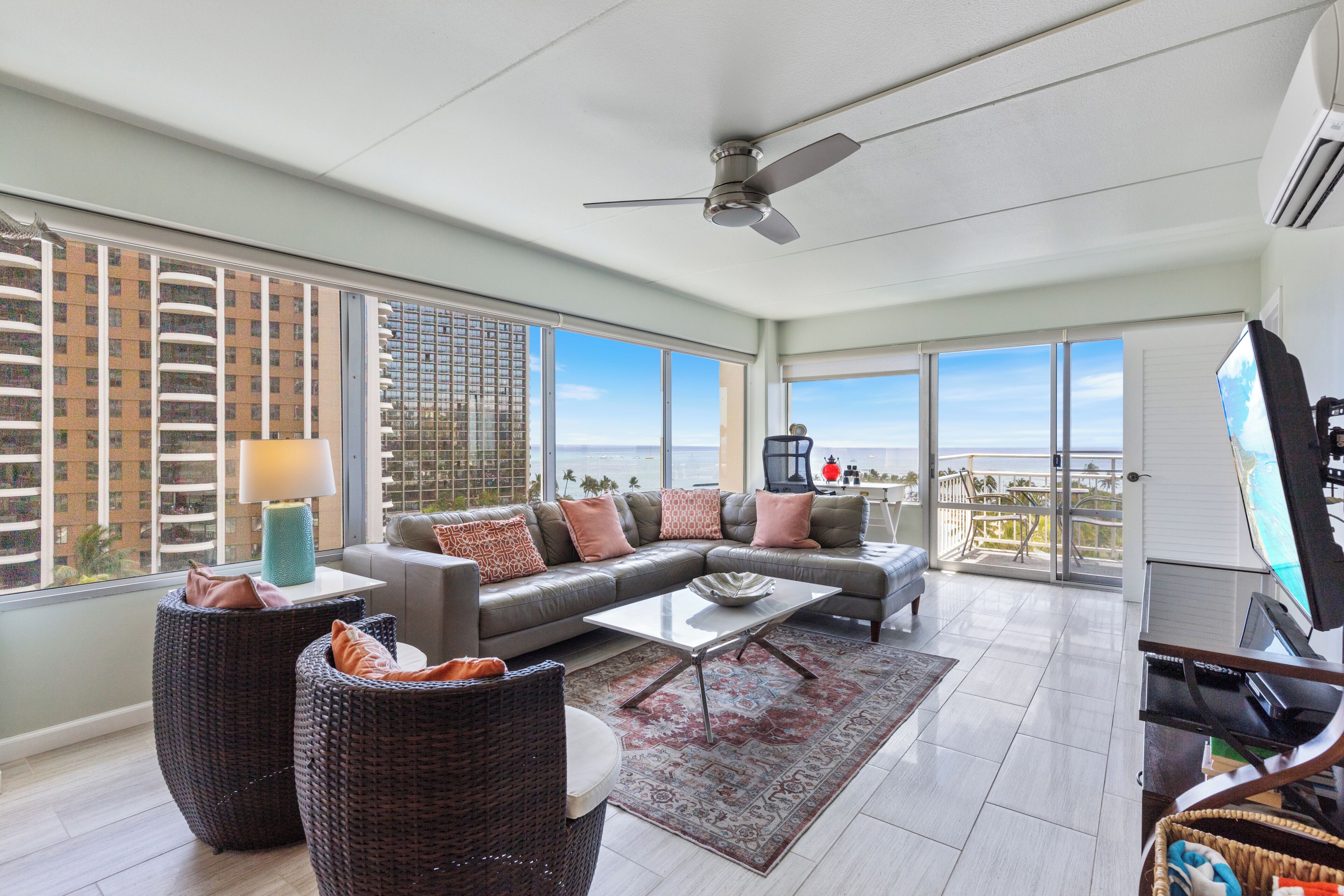 A spacious living room with a large sofa, a coffee table, and wicker chairs. There is a ceiling fan, a TV, and a view of the city through large windows.