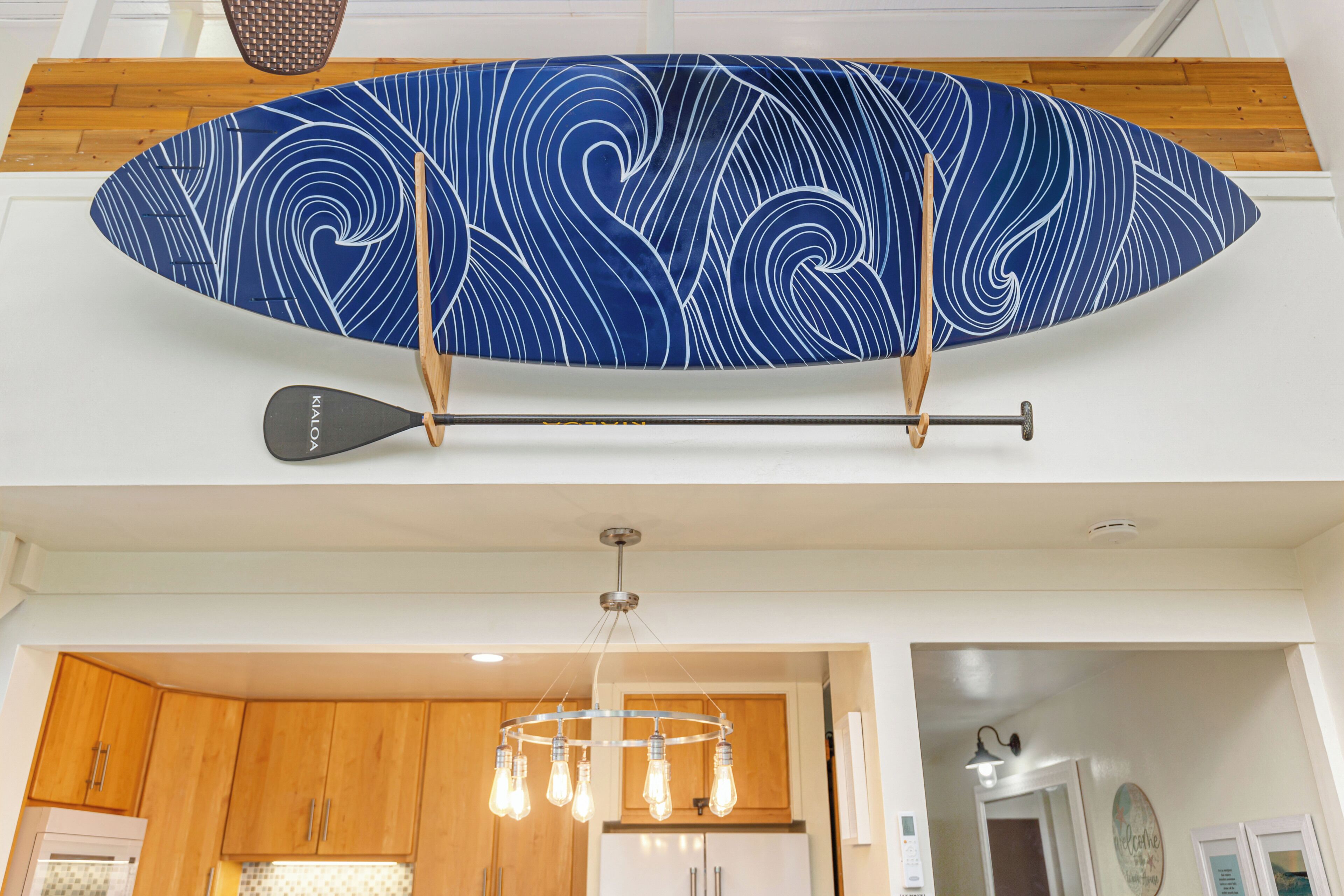 Un adorno mural con forma de tabla de surf, un remo, una cocina moderna con armarios de madera y una lámpara colgante.